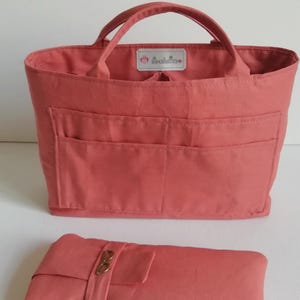 Könnte beinhalten: Ein korallenfarbener Handtaschen-Organizer aus Stoff mit zwei Tragegriffen, mehreren Außentaschen und einem Innenetikett mit dem Text "SOUBOUETTES". Darunter befindet sich eine passende korallenfarbene Stofftasche mit einem kleinen Brillen-Symbol.