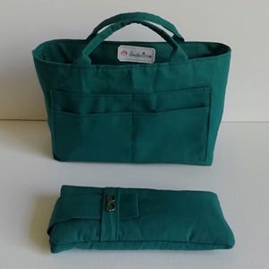Könnte beinhalten: Eine tealfarbene Tragetasche mit zwei Griffen und zwei Vordertaschen. Darunter befindet sich ein passender, rechteckiger, gefalteter Artikel in Teal mit einer goldfarbenen Schnalle. Die Tasche hat innen ein kleines weißes Etikett.