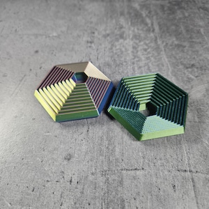 May include: Two hexagonal metal fidget spinners with a layered design. One spinner is green, blue, and yellow, and the other is yellow, green, and purple.