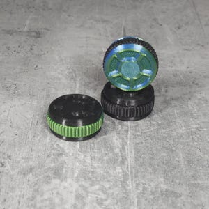 May include: Three 3D-printed bottle stoppers. Two are black with green and blue accents, and one is black with green accents. The stoppers have a ridged design around the sides and a circular top with a geometric pattern.