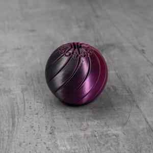 May include: A spherical object with a gradient color scheme, transitioning from dark gray to a vibrant purple. The surface features a series of curved, parallel lines, creating a textured appearance. The object is likely a decorative item or a puzzle.