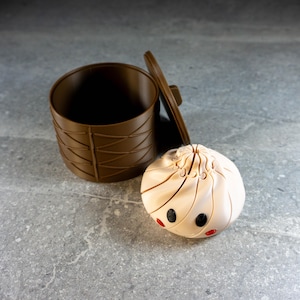 May include: A brown cylindrical container with a matching lid and a light beige, spherical, dumpling-shaped object featuring two black circular eyes and two small red circular accents on its lower front surface, positioned next to the container.
