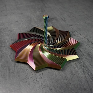May include: A multi-colored, geometric sculpture with a spiral stem. The sculpture features a star-like base with layered, angular segments in shades of red, gold, green, and purple. The stem is a twisted, striped design.