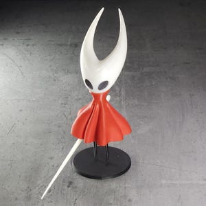 May include: A white and red figurine of a character with a curved headpiece, black eyes, and a red cape. The figurine is mounted on a black circular base and holds a white, slender weapon. The design is stylized.