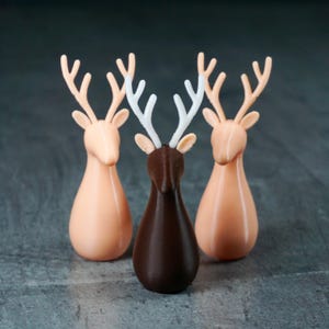May include: Three decorative deer figurines. Two are a light peach color, and one is dark brown. The antlers are a contrasting white or peach color. The deer have a smooth, rounded body shape and are standing upright.