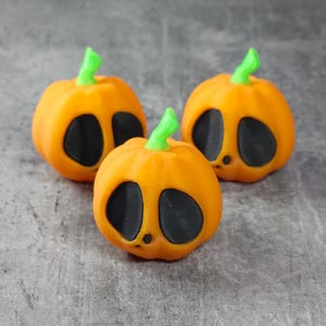 May include: Three orange pumpkin decorations with black eye and mouth cutouts and green stems. These small, decorative pumpkins are perfect for Halloween or fall-themed decor.