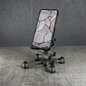 May include: A silver-colored phone stand with a tripod base supporting a smartphone. The phone displays a black and white circular pattern. The stand has adjustable joints and black rubber feet.