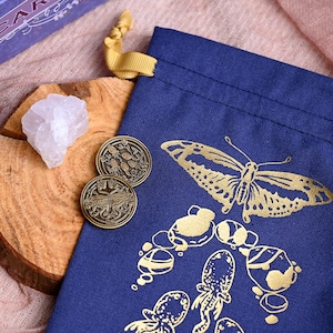 May include: A navy blue drawstring bag featuring a gold butterfly and octopus design. Two bronze coins with botanical and butterfly designs sit beside a clear quartz crystal on a wooden surface. A yellow ribbon is tied to the bag.