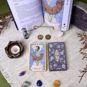 May include: An open book with an otter illustration, a tarot card with an otter illustration, and a deck of tarot cards with a floral design. Also, a candle holder, crystals, and coins are visible.