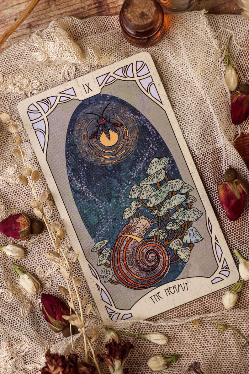 May include: A tarot card illustration depicting a snail with a mushroom-covered shell. Above, a moth flies near a glowing orb. The card is framed with decorative borders and the words "THE HERMIT".