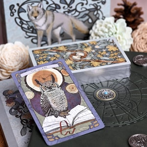 May include: A tarot card titled The Scribe featuring an owl perched on an open book, resting on a dark green textured surface next to a deck of cards with a botanical back design. The card has a purple border and a circular wood grain background.