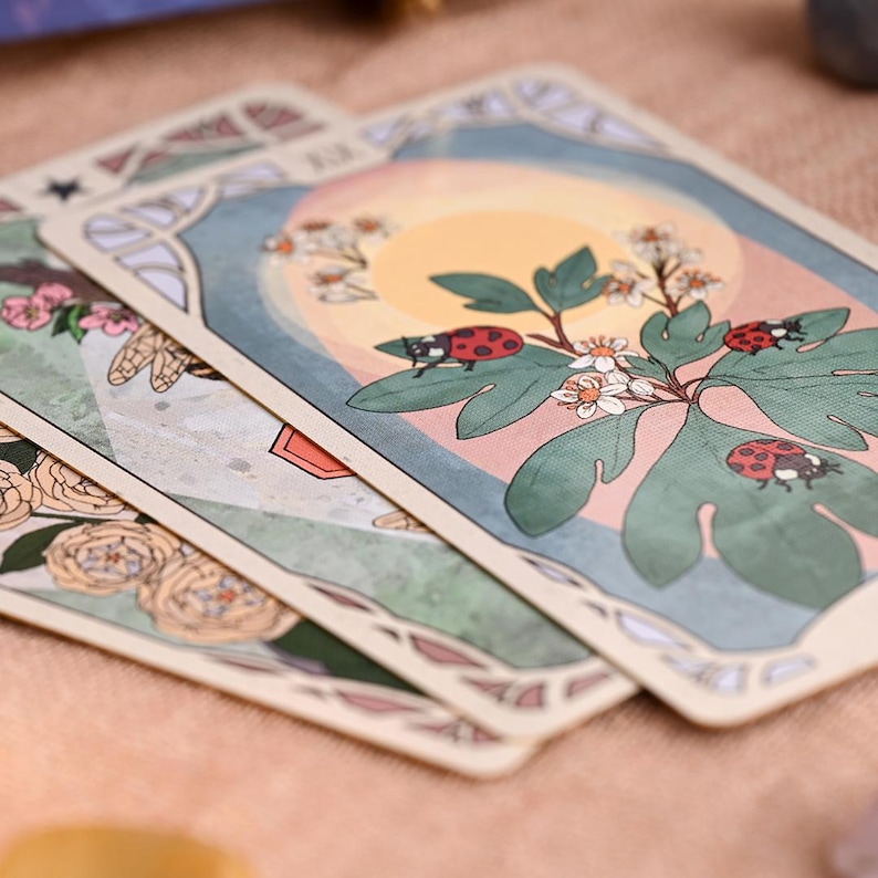 May include: Three tarot cards with illustrations of flowers and insects. One card shows a sun, leaves, and ladybirds. The other cards have floral and insect designs. The cards have a cream background and decorative borders.