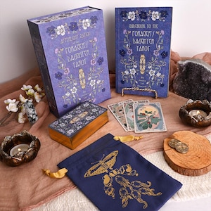 May include: A tarot card set featuring a purple box and guidebook, both decorated with floral patterns and the text "Afterlight Edition of the Goddess's Daughter Tarot." The set includes a card deck, a drawstring bag with a butterfly design, and decorative items.