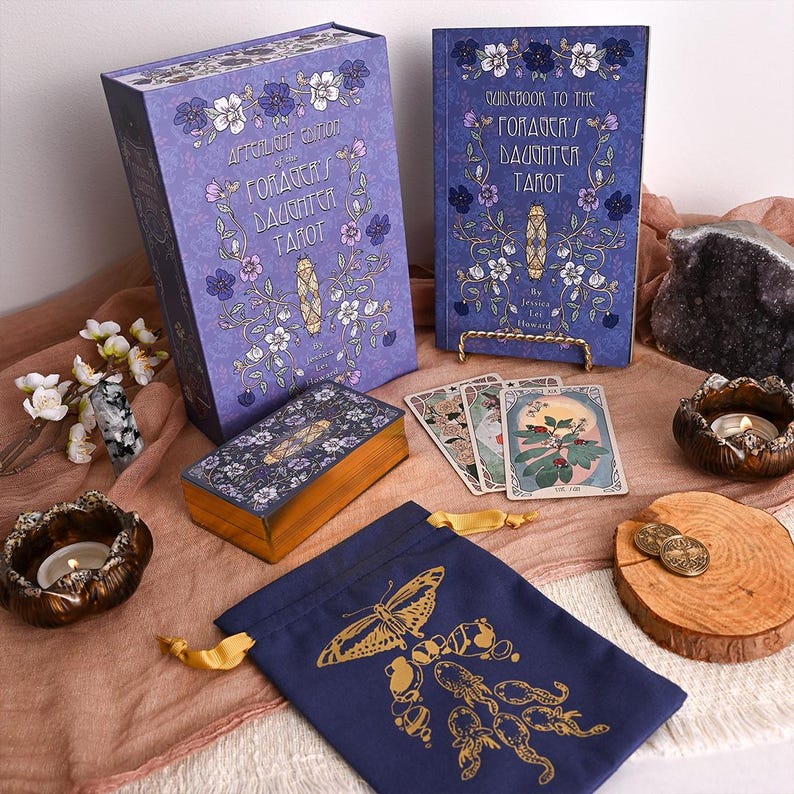 May include: A tarot card set featuring a purple box and guidebook, both decorated with floral patterns and the text "Afterlight Edition of the Goddess's Daughter Tarot." The set includes a card deck, a drawstring bag with a butterfly design, and decorative items.