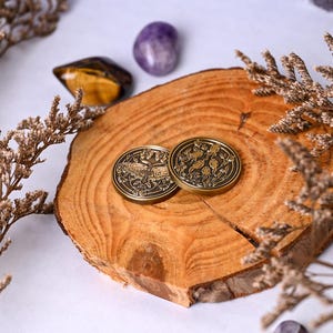 May include: Two antique bronze coins with detailed nature-themed designs rest on a wooden slice. One coin features a butterfly, the other a botanical pattern. Dried flowers and gemstones, including tiger's eye and amethyst, surround the coins.