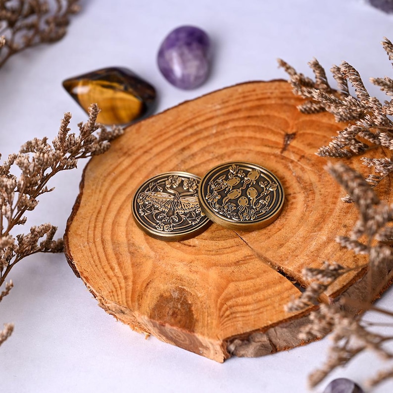 May include: Two antique bronze coins with detailed nature-themed designs rest on a wooden slice. One coin features a butterfly, the other a botanical pattern. Dried flowers and gemstones, including tiger's eye and amethyst, surround the coins.