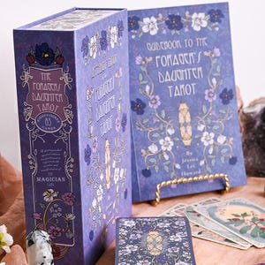 May include: A tarot card set with a purple box and guidebook. The box and guidebook feature floral designs and the text "The Gorda's Daughter Tarot." The set includes 83 cards, with the Magician card visible.
