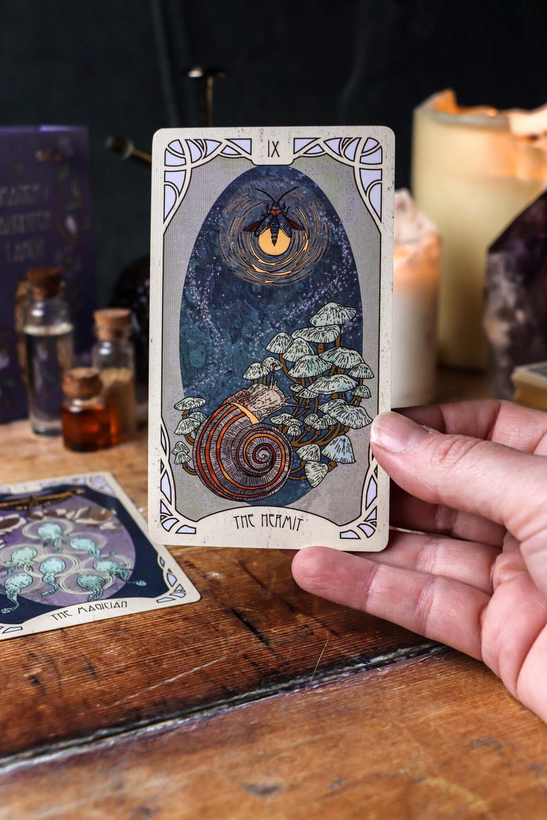 May include: A tarot card showing a snail with a mushroom-like growth, set against a dark blue backdrop with a glowing insect. The card is labelled "THE HERMIT" and is held in a hand.