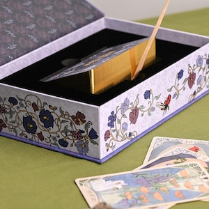 May include: A tarot card deck in a decorative box. The box is white with floral and vine designs in purple, blue, and red. The deck has gold edges and a gold ribbon. Several tarot cards are visible on the green surface.