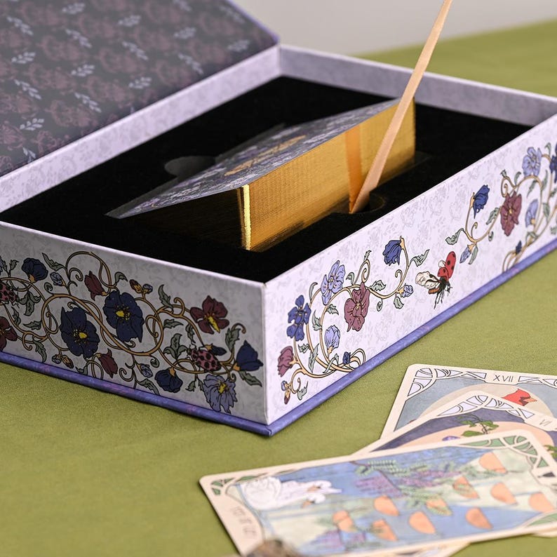 May include: A tarot card deck in a decorative box. The box is white with floral and vine designs in purple, blue, and red. The deck has gold edges and a gold ribbon. Several tarot cards are visible on the green surface.
