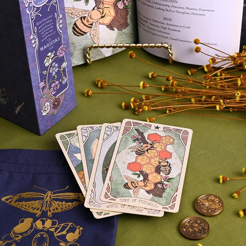 May include: A tarot card deck with a purple box featuring floral and gold accents. The cards display bee and honeycomb illustrations. A navy blue bag with a gold butterfly design and two gold coins are also visible.