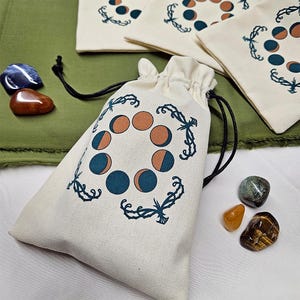 May include: A white cotton drawstring bag with a black and brown moon phase design. The bag is filled with various crystals and stones.