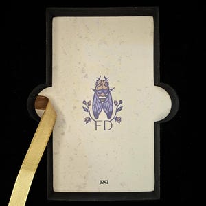 May include: A rectangular card with a black border and a gold ribbon. The card features a fly illustration with the initials "FD" and the number "0242". The background is off-white with a speckled texture.