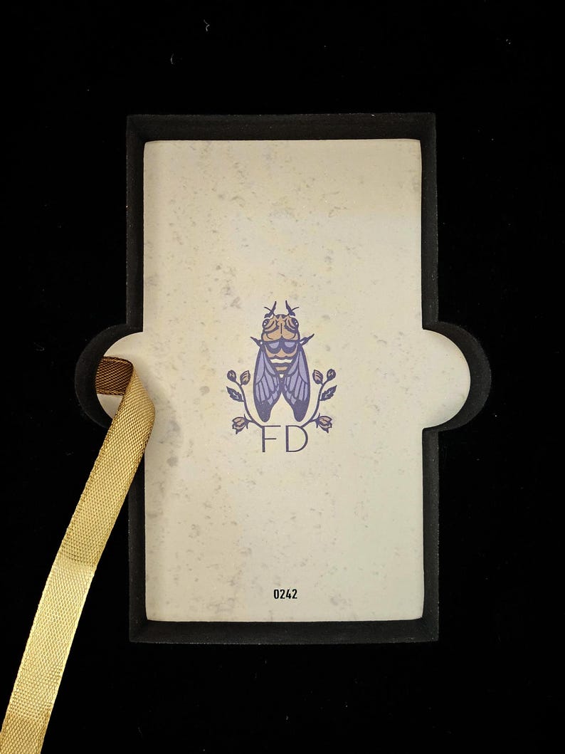 May include: A rectangular card with a black border and a gold ribbon. The card features a fly illustration with the initials "FD" and the number "0242". The background is off-white with a speckled texture.
