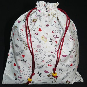 Peut inclure: Sac cadeau en tissu blanc avec un imprimé sur le thème de Noël représentant des baies rouges, des nœuds dorés et des feuilles grises. Le sac a des cordons de serrage rouges avec des perles jaunes.