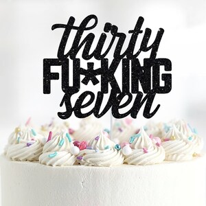 May include: Black glitter cake topper with the text "thirty f*cking seven" on a white frosted cake with colourful sprinkles.
