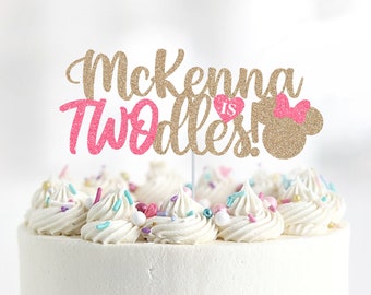 Può includere: Topper per torta in glitter dorato con il testo "McKenna is Two-dles!" in glitter rosa e dorato. Il topper è decorato con un fiocco rosa e una testa di topo in glitter dorato.