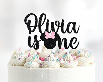 May include: Black glitter cake topper with the text "Olivia is one" and a black and pink glitter Minnie Mouse head.