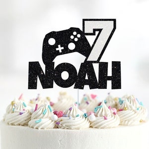 Peut inclure: Un topper de gâteau noir pailleté avec une manette de jeu vidéo, le chiffre 7 et le nom Noah. Le topper est sur un gâteau blanc avec des vermicelles colorés.