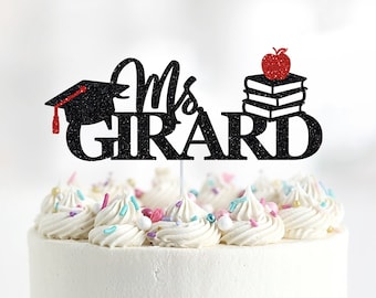 May include: Black glitter cake topper with the text "Mrs. Girard" and a graduation cap, a stack of books, and a red apple.