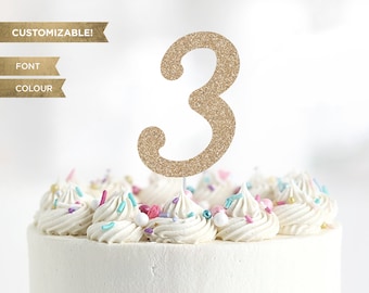 May include: Gold glitter number three cake topper. Customizable font and color. The topper is on a white cake with colorful sprinkles.