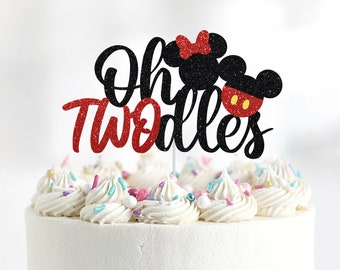 May include: A black and red cake topper with the text "Oh Twodles" and a cartoon image of Mickey and Minnie Mouse. The topper is on top of a white frosted cake with colorful sprinkles.