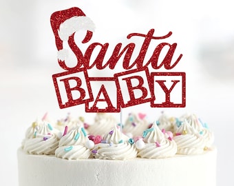May include: Red glitter cake topper with a Santa hat and the words "Santa Baby" in block letters. The topper is on a white frosted cake with sprinkles.