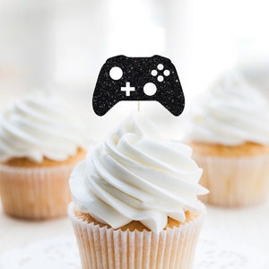 May include: Three cupcakes with white frosting. The cupcake in the center has a black glitter game controller topper.