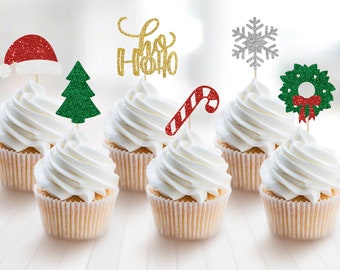 May include: Set of six white cupcakes with various Christmas themed toppers. The toppers include a red Santa hat, a green Christmas tree, a gold glitter "Ho Ho Ho" sign, a candy cane, a silver snowflake, and a green wreath with a red bow.