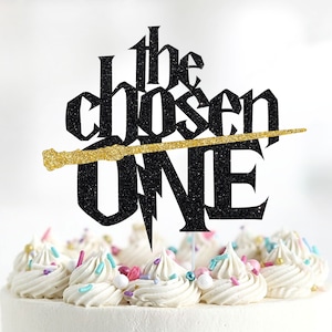 May include: Black glitter cake topper with the text "the chosen one" and a gold glitter wand. The wand is positioned diagonally across the text.