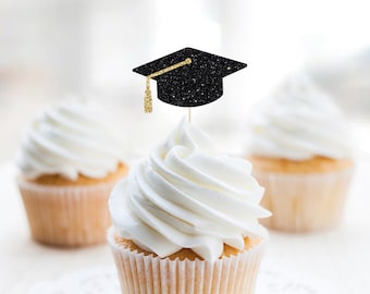 May include: Three white cupcakes with black glitter graduation cap toppers. The cupcakes are on a white surface.