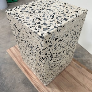 Könnte beinhalten: Ein quadratischer Betonschemel mit einer weißen Basis und schwarzen Flecken. Der Schemel steht auf einer Holzoberfläche.
