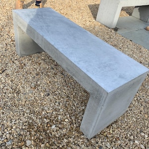 May include: Two concrete benches with a modern, minimalist design. The benches are made of light grey concrete and have a smooth, polished finish. The benches are sitting on a bed of gravel.