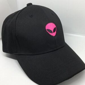 May include: Black baseball cap with a curved brim. The cap features a pink embroidered alien head on the front. The cap is made of a durable fabric and has a structured fit.
