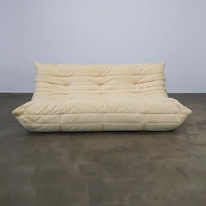 May include: A cream-coloured, low-profile sofa with a quilted design. The sofa has a rounded shape and a series of horizontal seams across the back and seat. The cushions are button-tufted. The sofa is set against a plain white wall.