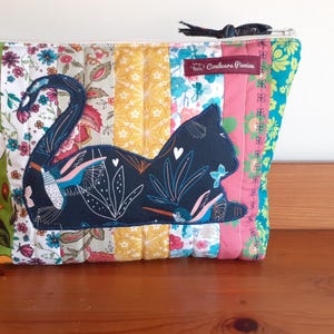 May include: A quilted pouch with a patchwork design featuring a black cat silhouette. The pouch has a zipper closure and is adorned with floral and patterned fabrics in various colors, including green, pink, and yellow. The text "Couleurs Pocine" is on a small label.