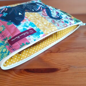 May include: A colorful, quilted pouch with a zipper closure. The patchwork design features floral and geometric patterns in pink, blue, yellow, and green. A dark blue cat silhouette is appliqued on the top. The interior is yellow with white polka dots.