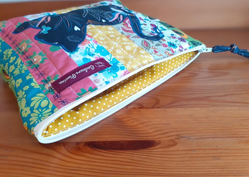 May include: A colorful, quilted pouch with a zipper closure. The patchwork design features floral and geometric patterns in pink, blue, yellow, and green. A dark blue cat silhouette is appliqued on the top. The interior is yellow with white polka dots.
