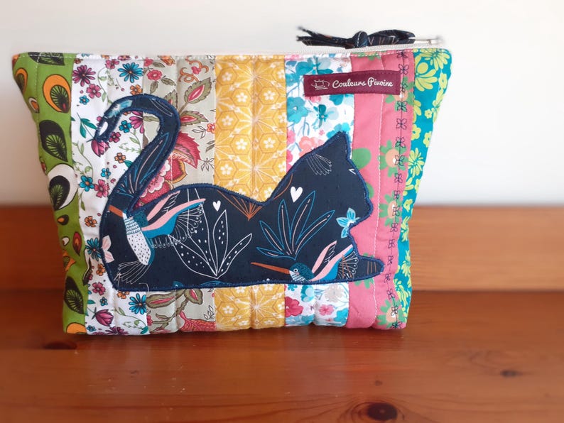 May include: A quilted pouch with a patchwork design featuring a dark blue cat silhouette appliqu&eacute;. The pouch has a zipper closure and a small tag that reads "Couleurs Pivoine". The patchwork includes floral and geometric patterns in various colors.