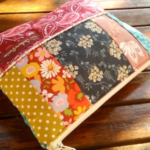 Peut inclure: Une pochette rectangulaire en patchwork avec une fermeture éclair. La pochette présente une variété de tissus aux couleurs bordeaux, jaune, orange et sarcelle. Les tissus comprennent des motifs floraux, cachemire et à pois. L'étiquette indique "Couleurs Poussière".